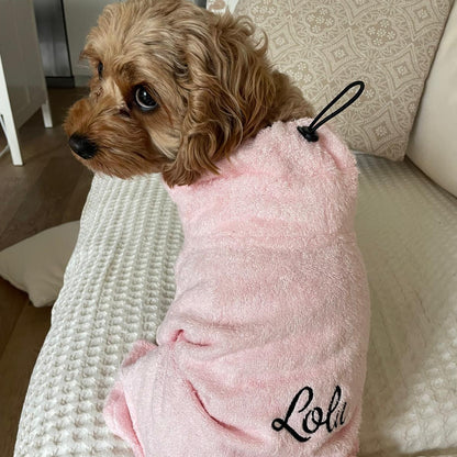 A small brown dog sitting on a cream textured sofa, wearing a plush light pink pet robe with the name 'Lola' embroidered in black script on the back.