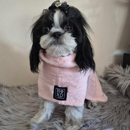 A small black and white dog wearing a pink plush pet cape with a black square patch that reads 'BxD' on the front, sitting on a gray shag rug.